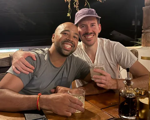 Mitchell and Takashi smiling with drinks at a patio table at night, one with an arm around the other